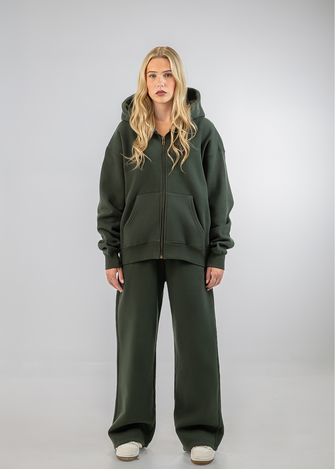 A woman modeling an oversized dark olive green zip-up hoodie and matching wide-leg sweatpants against a light gray studio background.