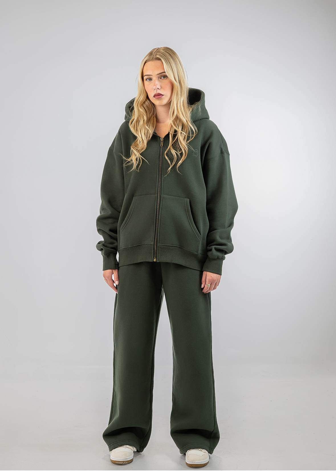 A blonde woman stands against a light gray studio background wearing an oversized dark olive green zip-up hoodie and matching wide-leg sweatpants.