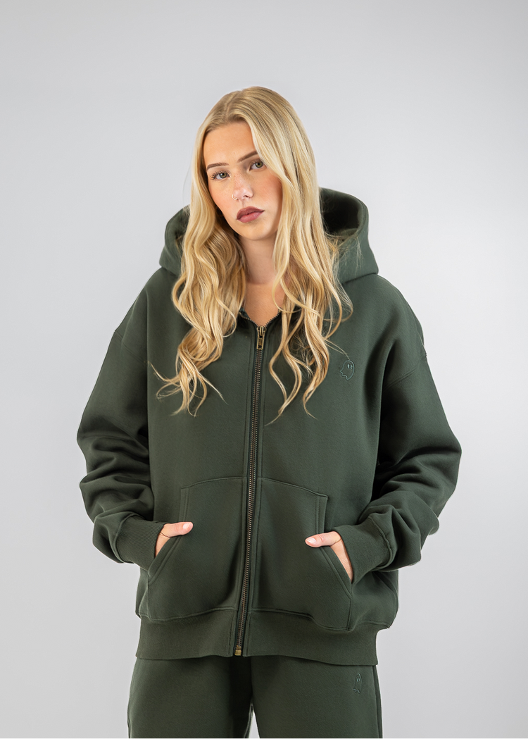 A blonde woman wearing an olive green zip-up hoodie and matching sweatpants, standing against a neutral gray studio background.