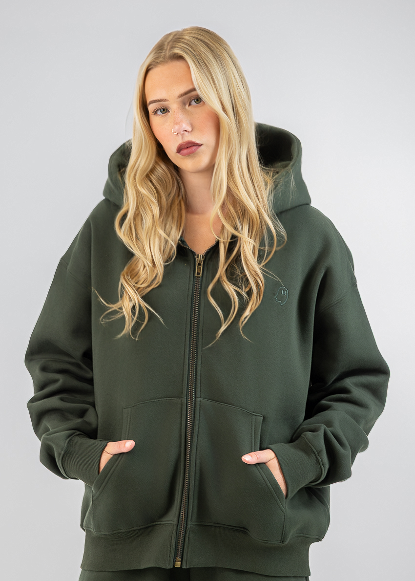 A blonde woman wearing an olive green zip-up hoodie and matching sweatpants, standing against a neutral gray studio background.