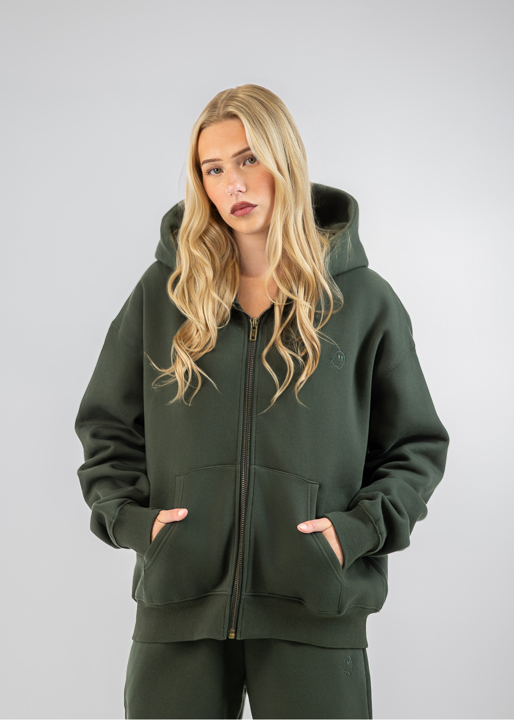 A blonde woman wearing an olive green zip-up hoodie and matching sweatpants, standing against a neutral gray studio background.