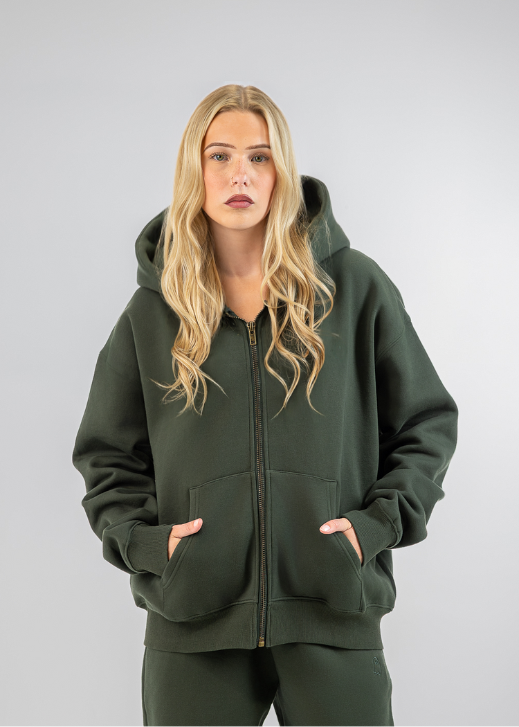 A blonde woman modeling an oversized dark green zip-up hoodie and matching sweatpants against a light gray studio background.
