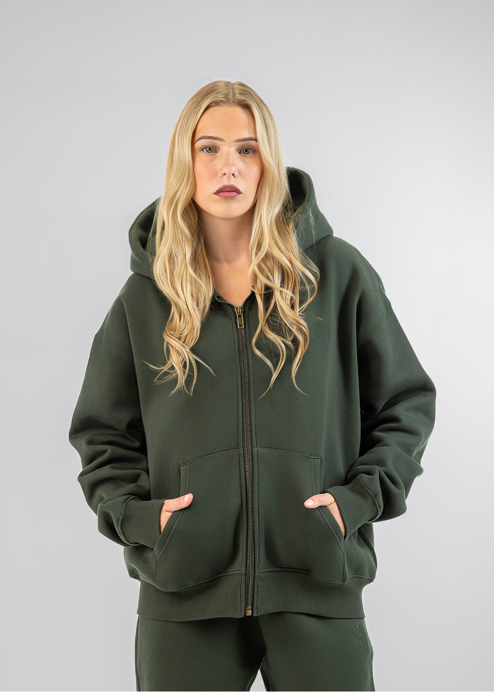 A blonde woman modeling an oversized dark green zip-up hoodie and matching sweatpants against a light gray studio background.