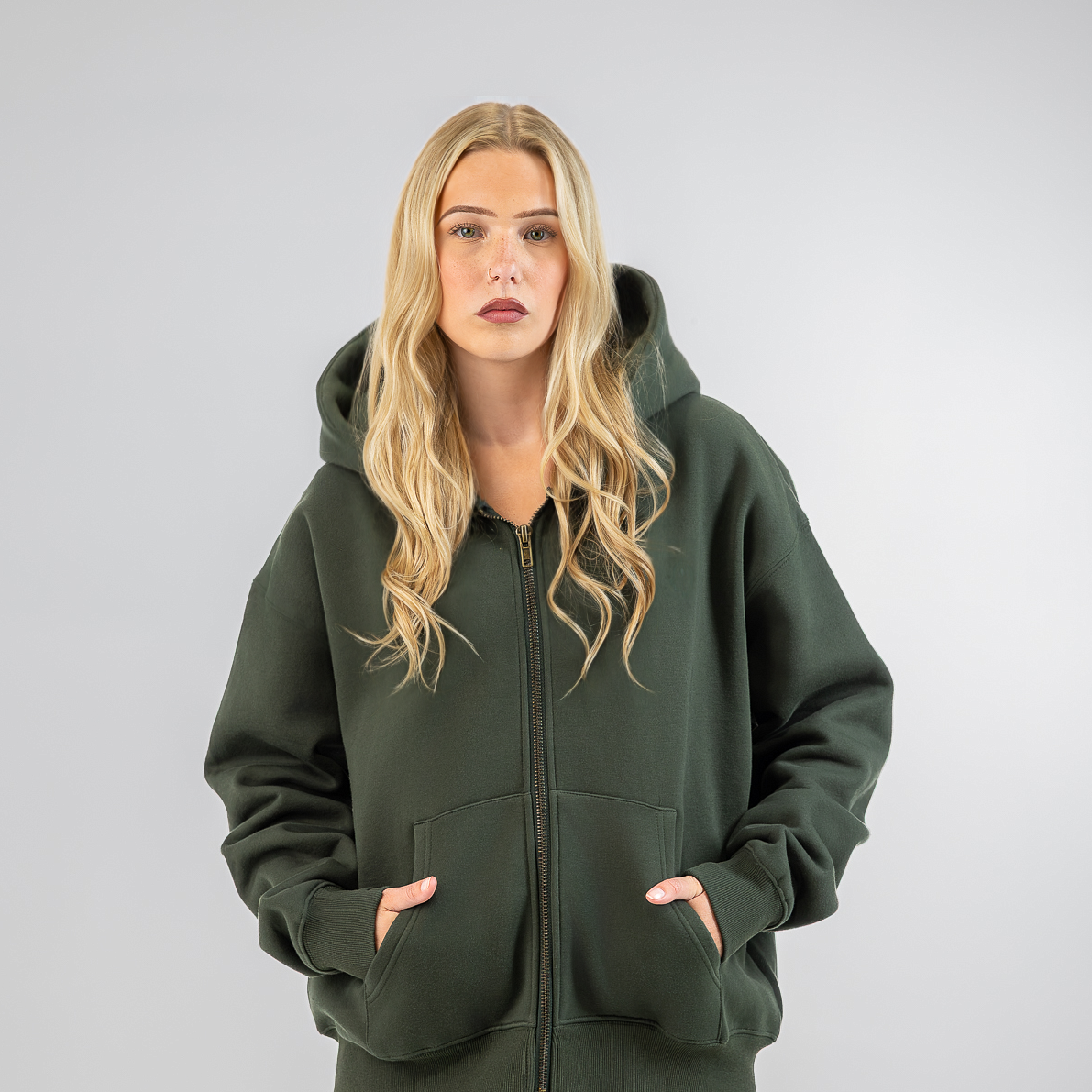 A blonde woman modeling an oversized dark green zip-up hoodie and matching sweatpants against a light gray studio background.