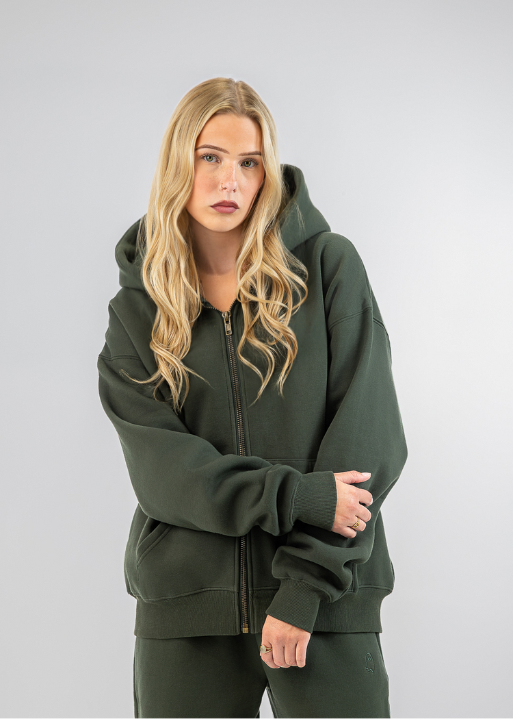 A blonde woman modeling a dark olive green zip-up hoodie and matching sweatpants against a light gray studio background.