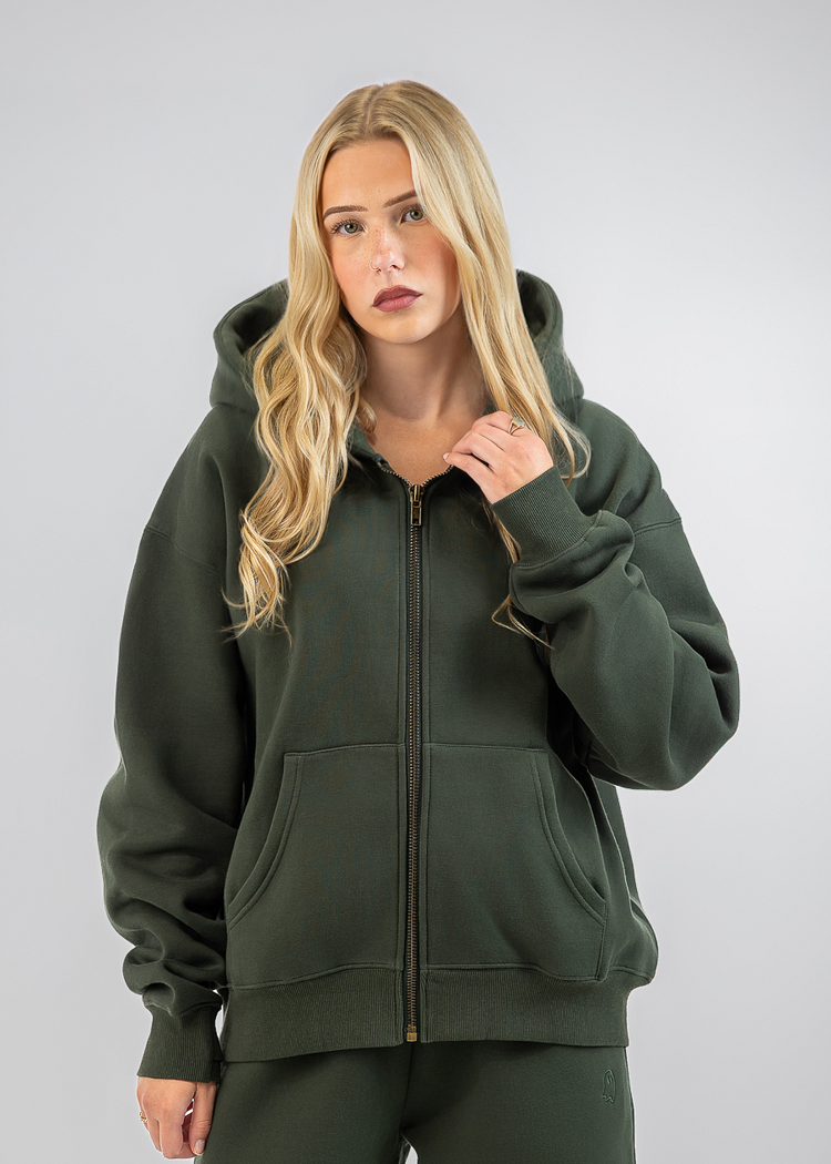 A blonde woman modeling a dark olive green zip-up hoodie and matching sweatpants against a light gray studio background.