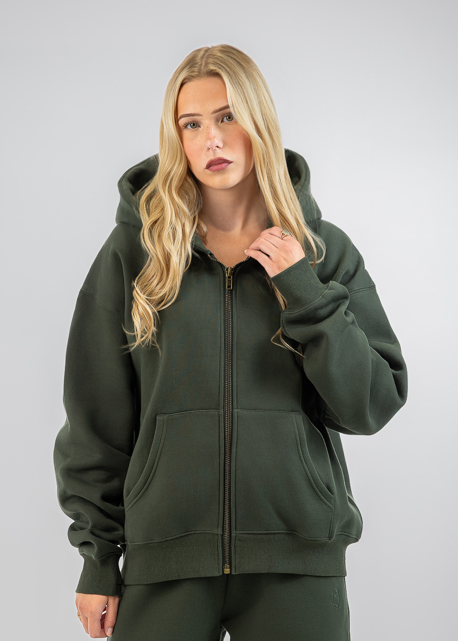 A blonde woman modeling a dark olive green zip-up hoodie and matching sweatpants against a light gray studio background.