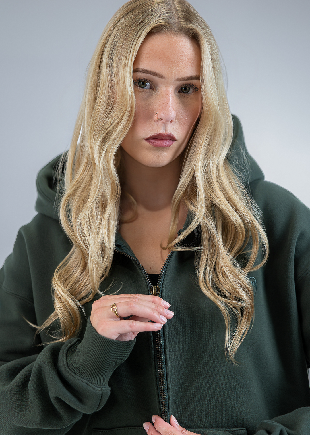 A blonde woman wearing an oversized dark green zip-up hoodie, posing against a neutral gray studio background.