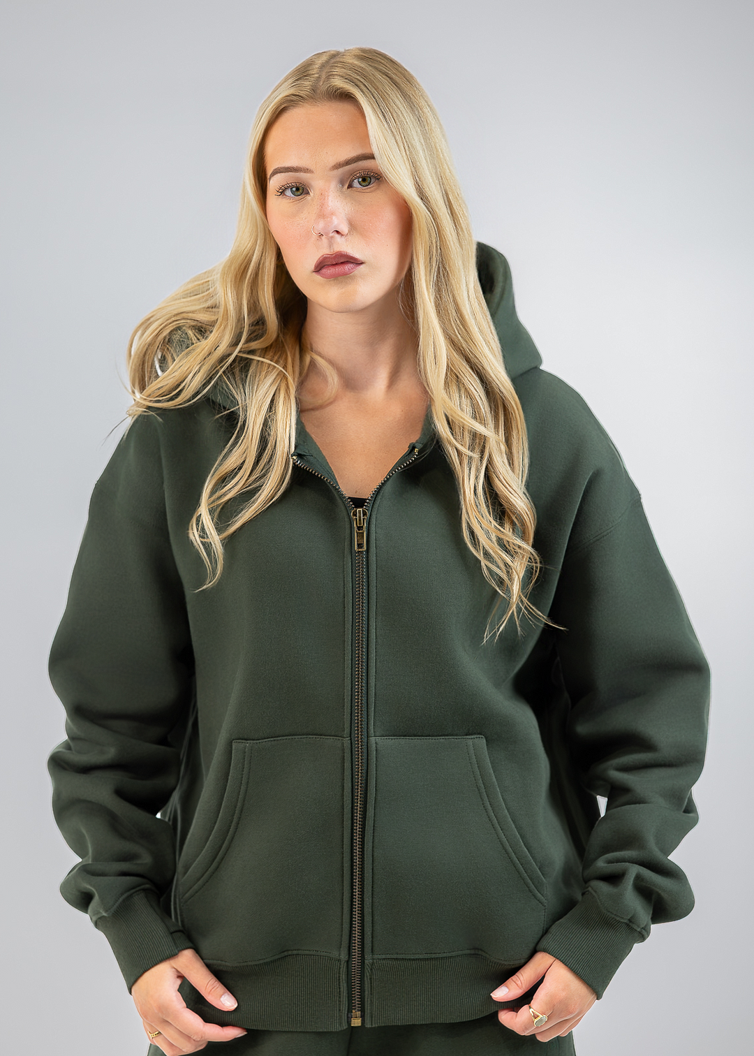 A blonde woman wearing a dark olive green zip-up hoodie with a front kangaroo pocket, standing against a neutral gray studio background.