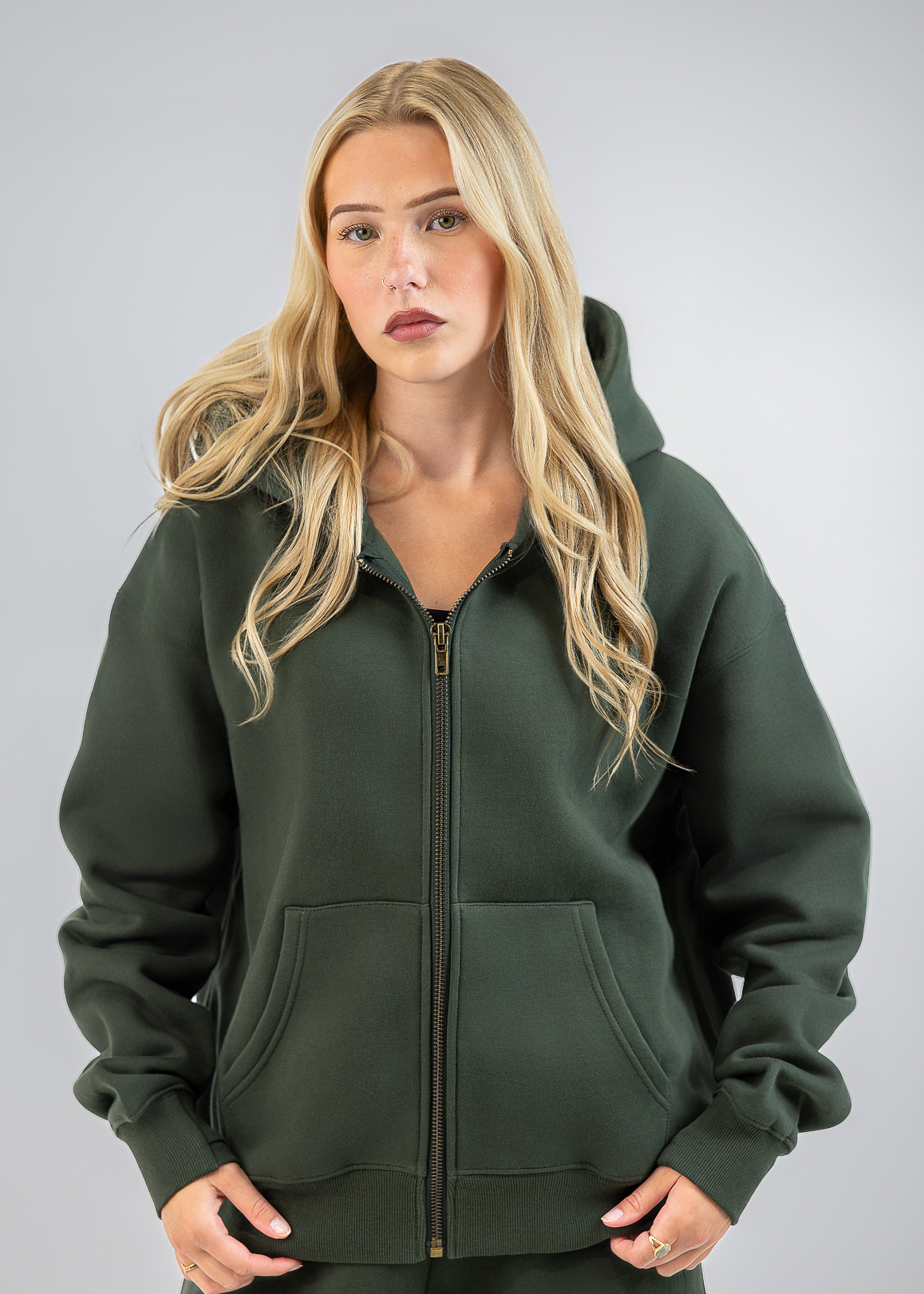 A blonde woman wearing a dark olive green zip-up hoodie with a front kangaroo pocket, standing against a neutral gray studio background.