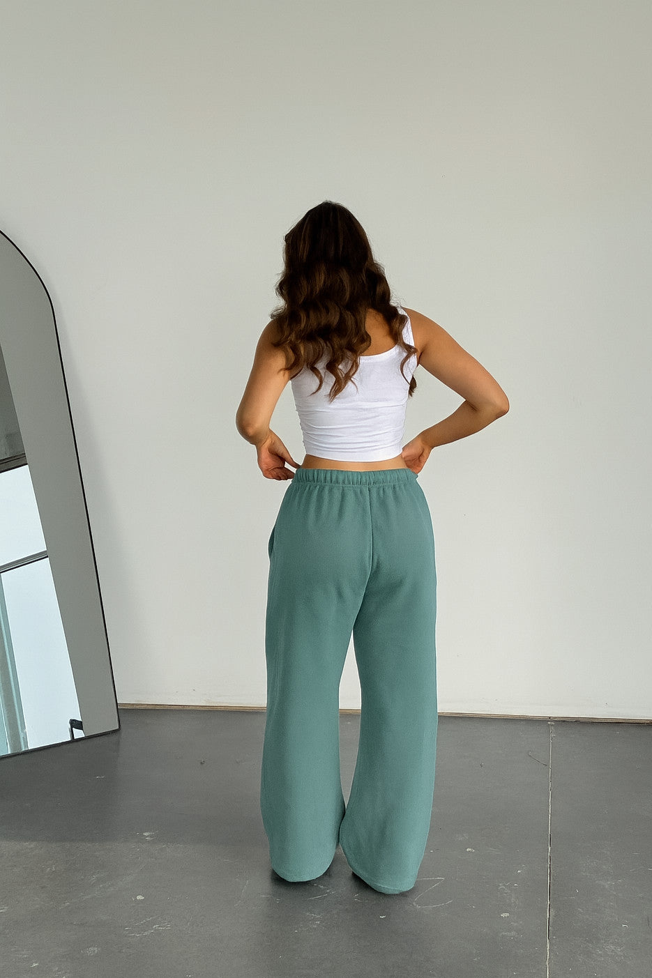 Woman wearing a white top and green pants standing in front of a mirror.