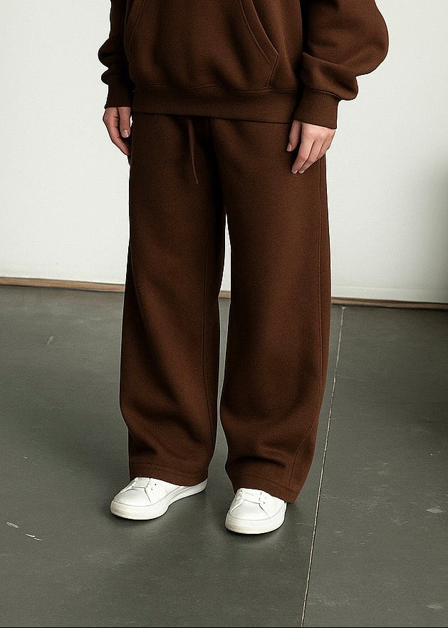 Person wearing a brown hoodie and pants standing in a minimalistic room.