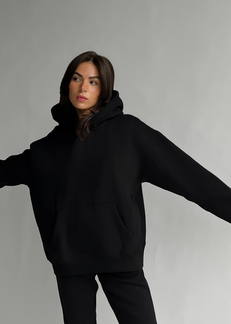 A woman modeling an oversized black pullover hoodie with a front kangaroo pocket and matching black pants, posed against a plain light gray studio background.