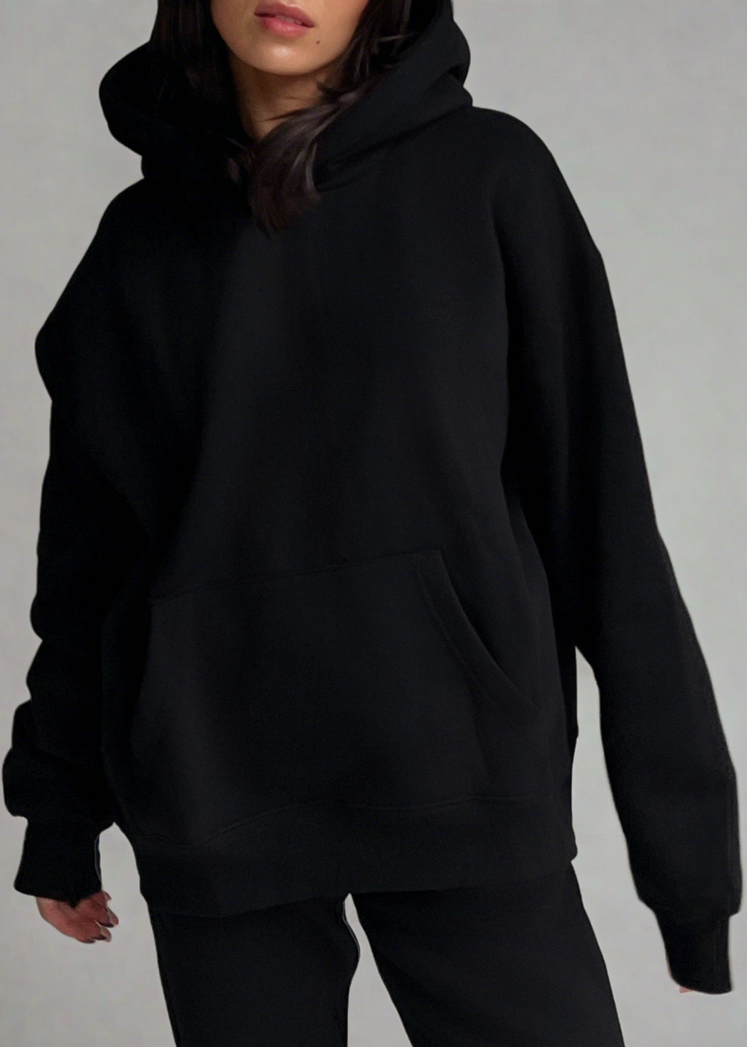 A person wearing an oversized black pullover hoodie with a front kangaroo pocket, paired with black pants, photographed against a plain light gray studio background.