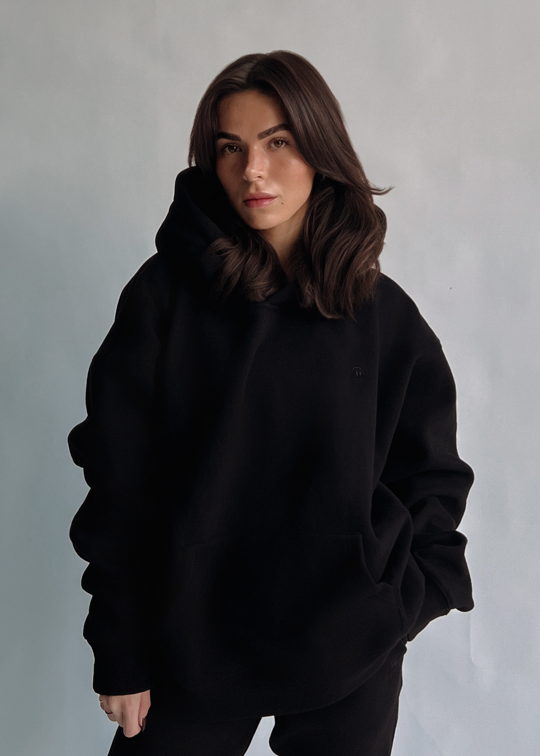 A woman with shoulder-length brown hair wearing an oversized black pullover hoodie and black pants, standing against a textured light gray studio background.