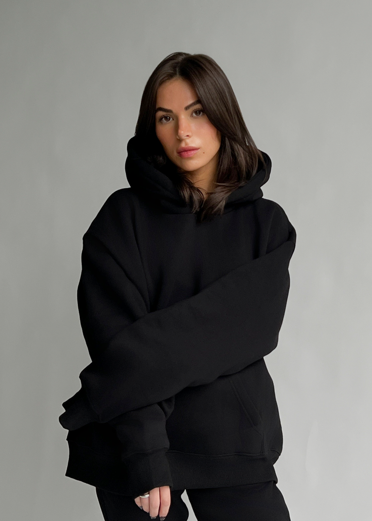 A woman wearing an oversized black pullover hoodie with a high collar and long sleeves, posed against a neutral gray studio background.