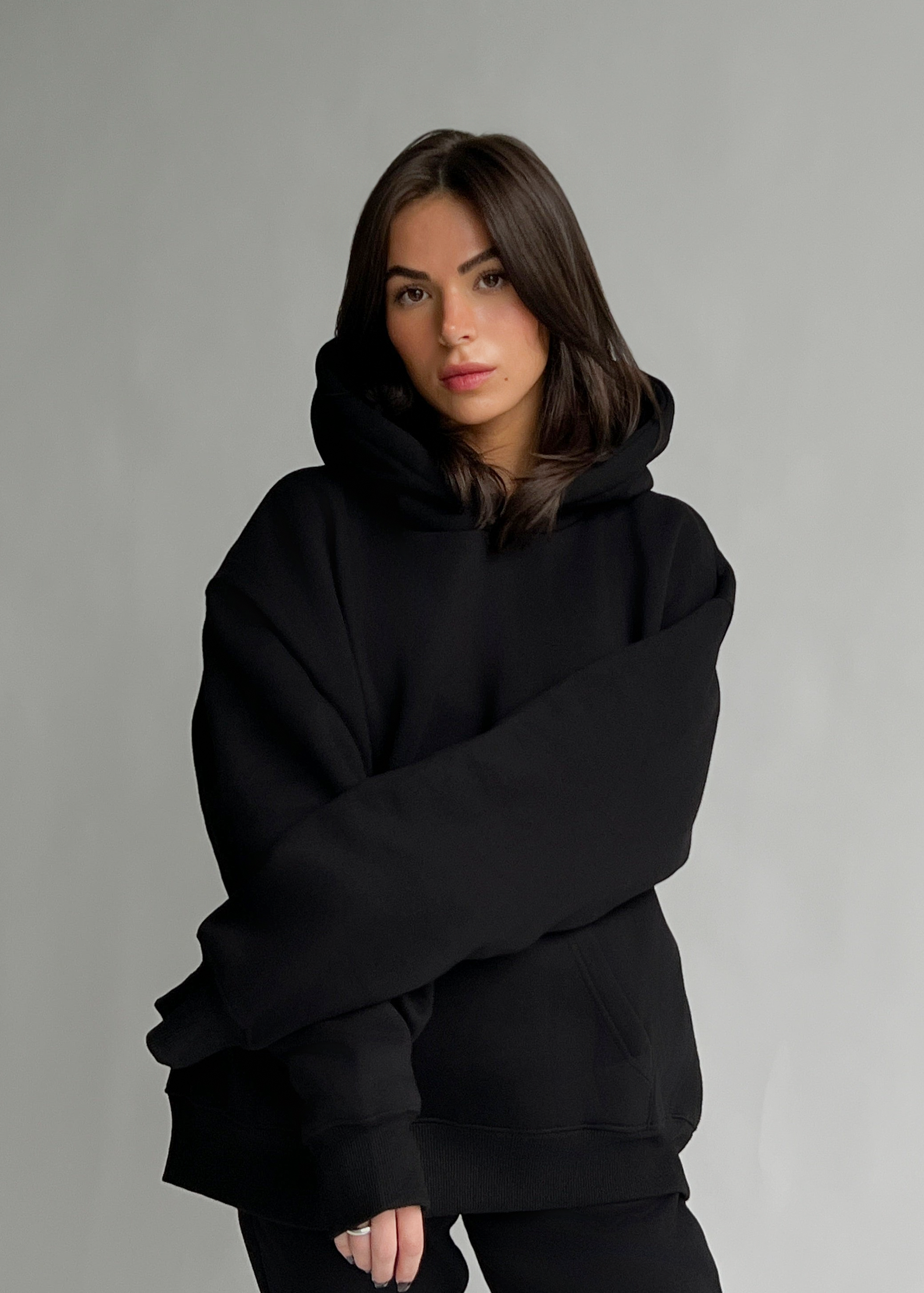 A woman wearing an oversized black pullover hoodie with a high collar and long sleeves, posed against a neutral gray studio background.