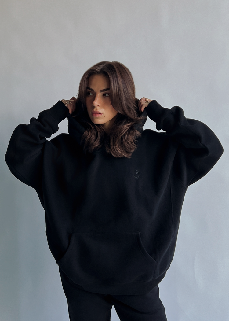 A woman with shoulder-length brown hair wearing an oversized black hoodie and matching black pants, posing with her hands behind her head against a plain light gray wall.