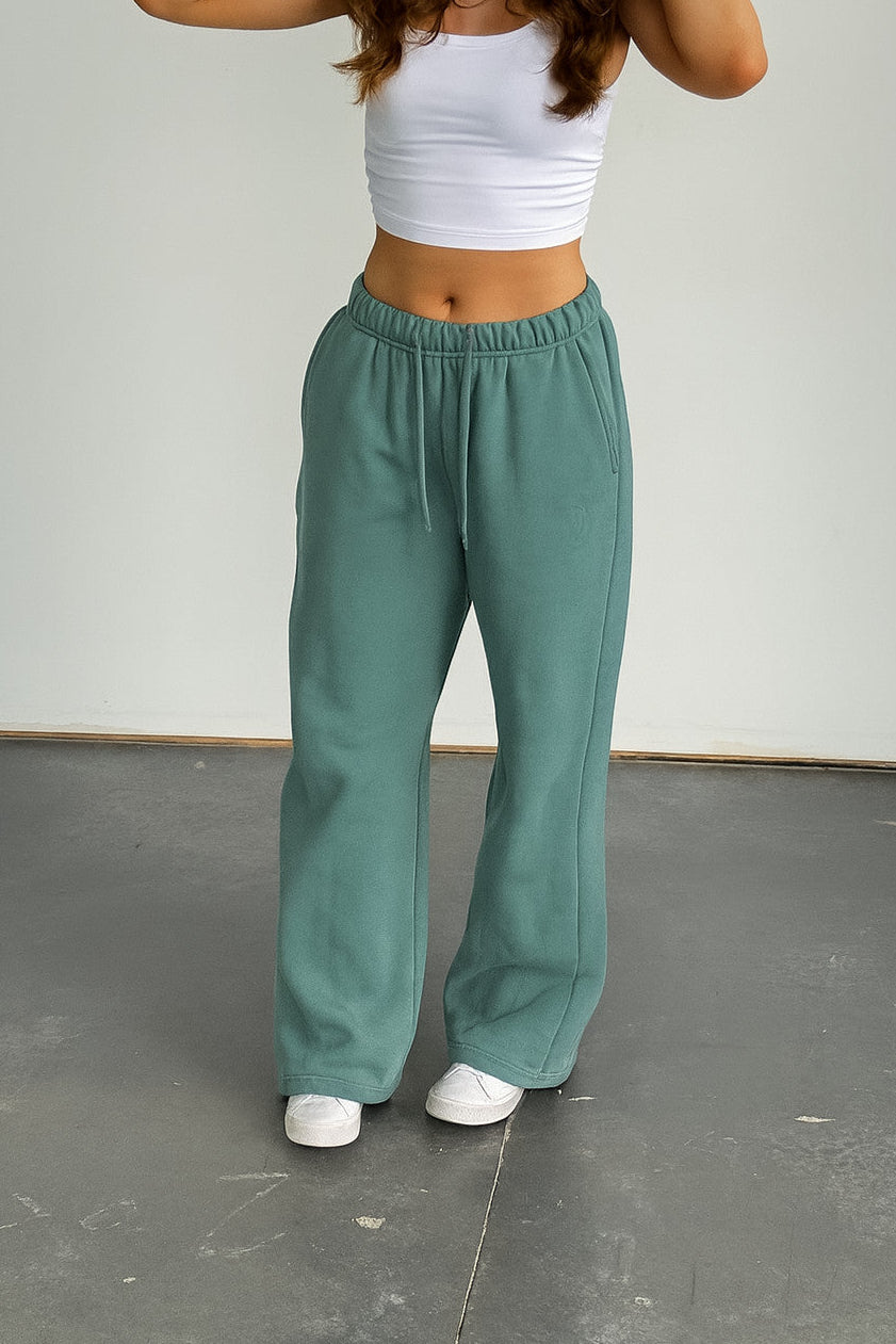 Person wearing teal pants and a white top on a gray floor with a white wall background