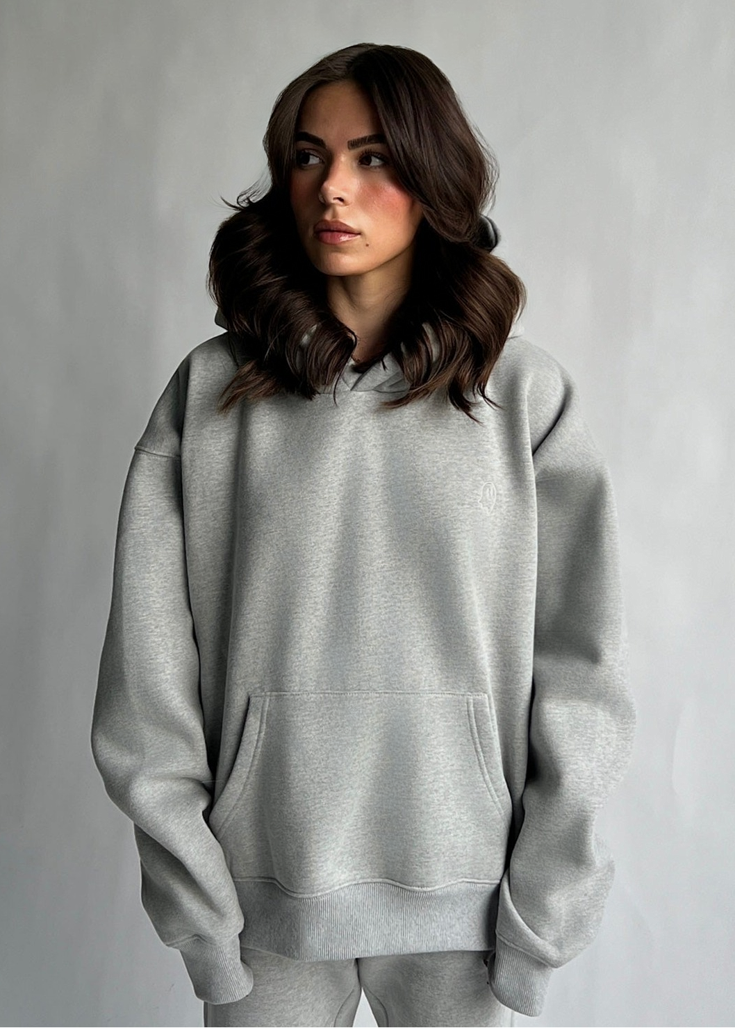 A woman with wavy brown hair wearing a light gray oversized pullover hoodie and matching gray sweatpants, standing against a neutral gray studio background.