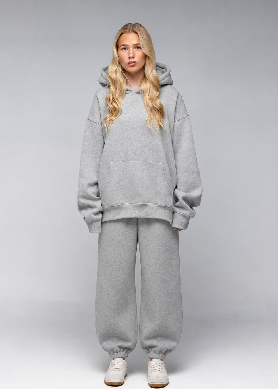 A blonde model stands against a light gray studio background wearing a matching light gray oversized hoodie and jogger sweatpants set, paired with white sneakers.