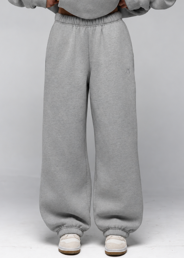 A person wearing light gray sweatpants and a matching gray sweatshirt, standing against a light gray studio background. The photo is cropped from the chest down, showing the full length of the sweatpants and white sneakers.
