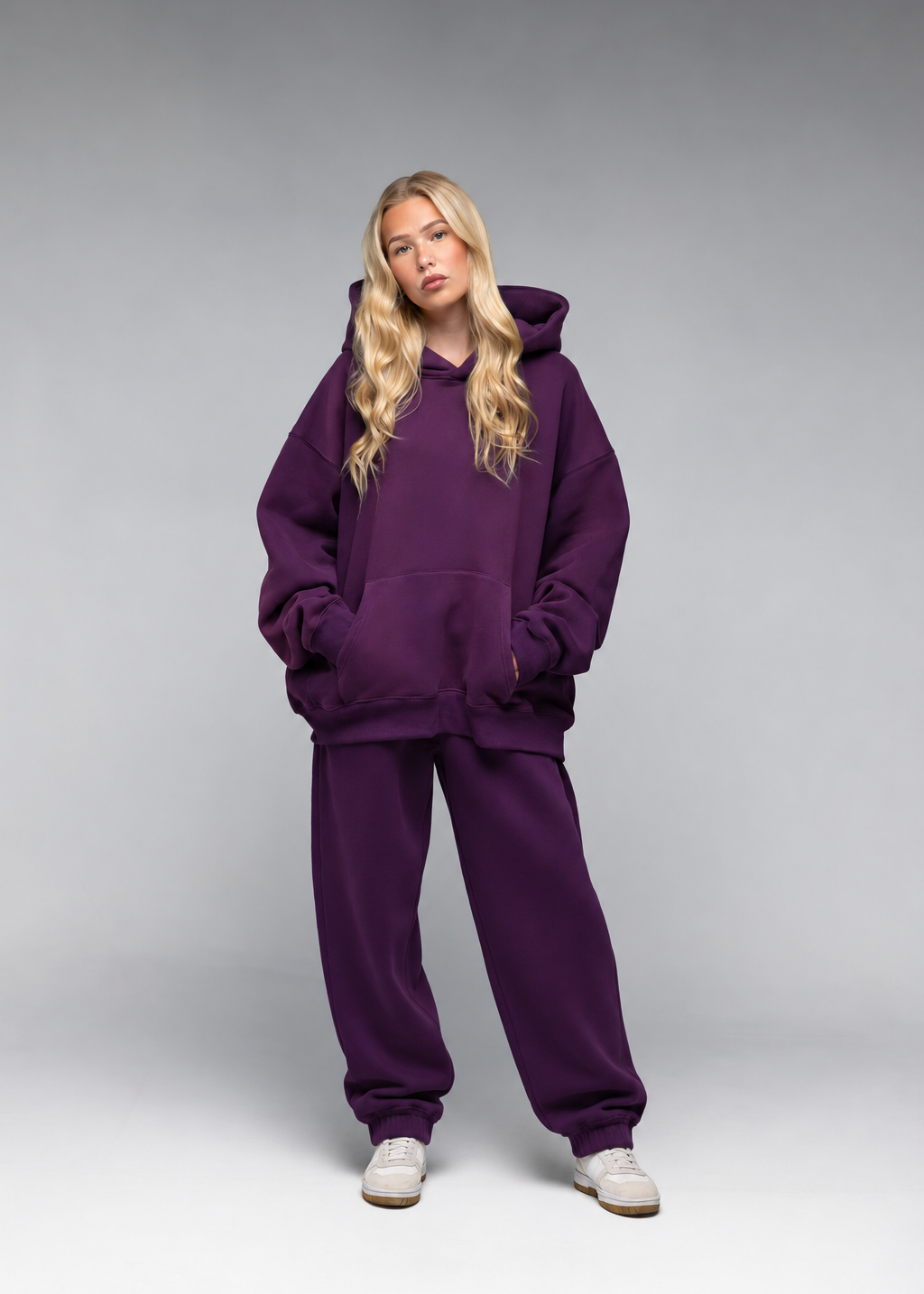 A blonde woman modeling an oversized deep purple hoodie and matching sweatpants set, paired with white sneakers, against a neutral gray studio background.