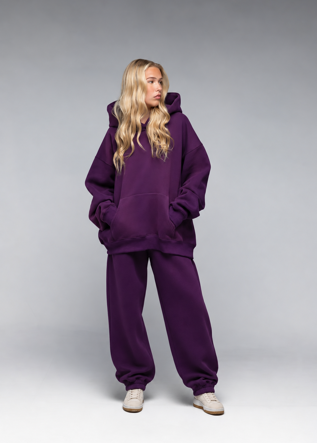 A blonde model wearing an oversized deep purple hoodie and matching sweatpants, standing against a neutral gray studio background.