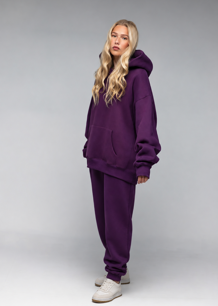 A blonde model wearing a matching deep purple sweatshirt and jogger pants set, paired with white sneakers, standing against a neutral gray studio background.