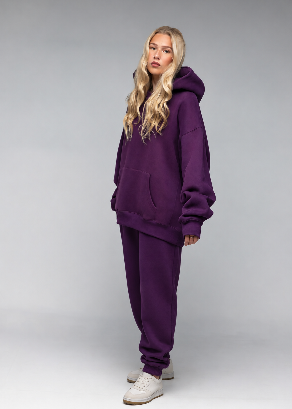 A blonde model wearing a matching deep purple sweatshirt and jogger pants set, paired with white sneakers, standing against a neutral gray studio background.