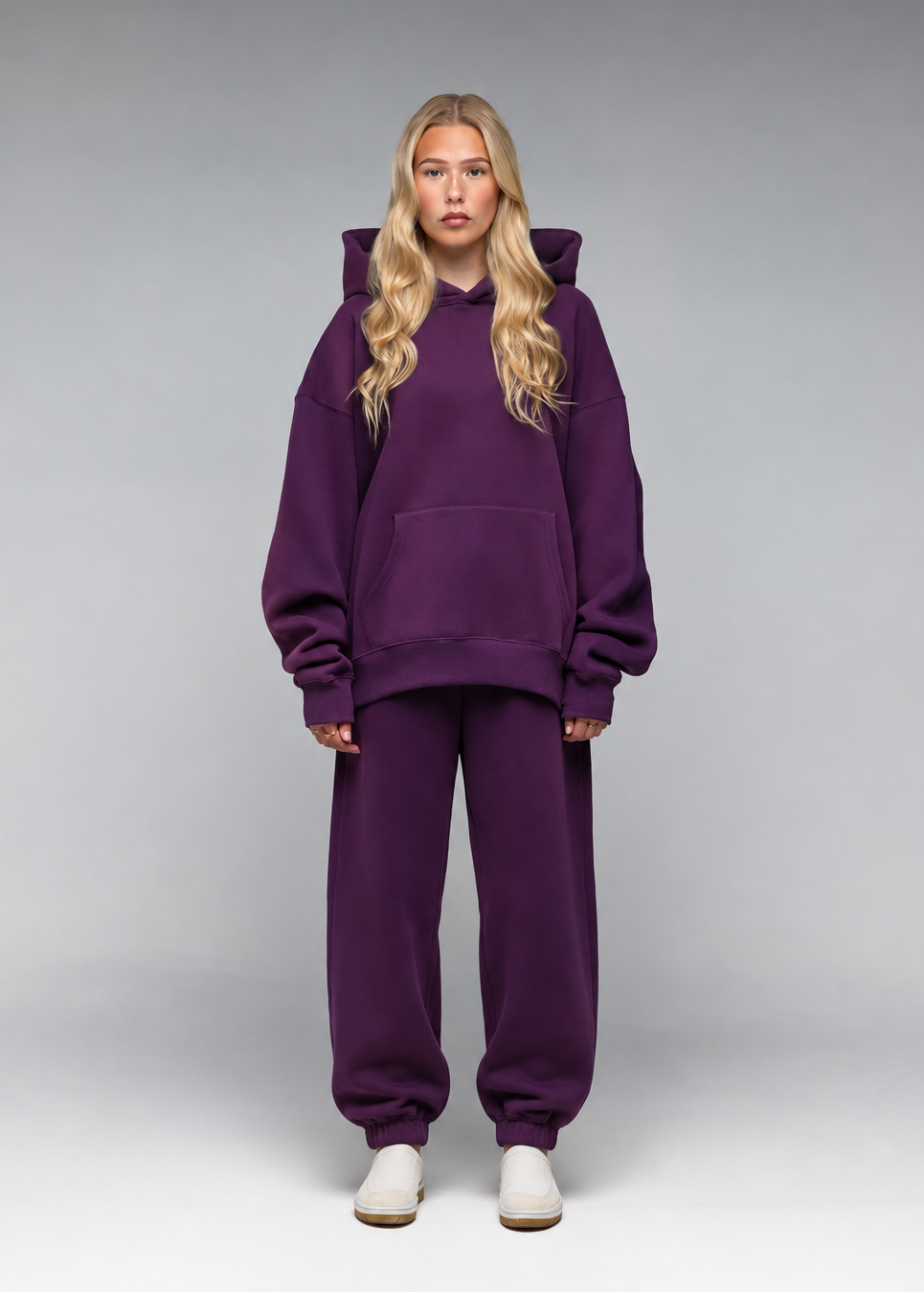 A blonde woman stands against a gray studio background wearing an oversized matching purple hoodie and sweatpants set with white sneakers.