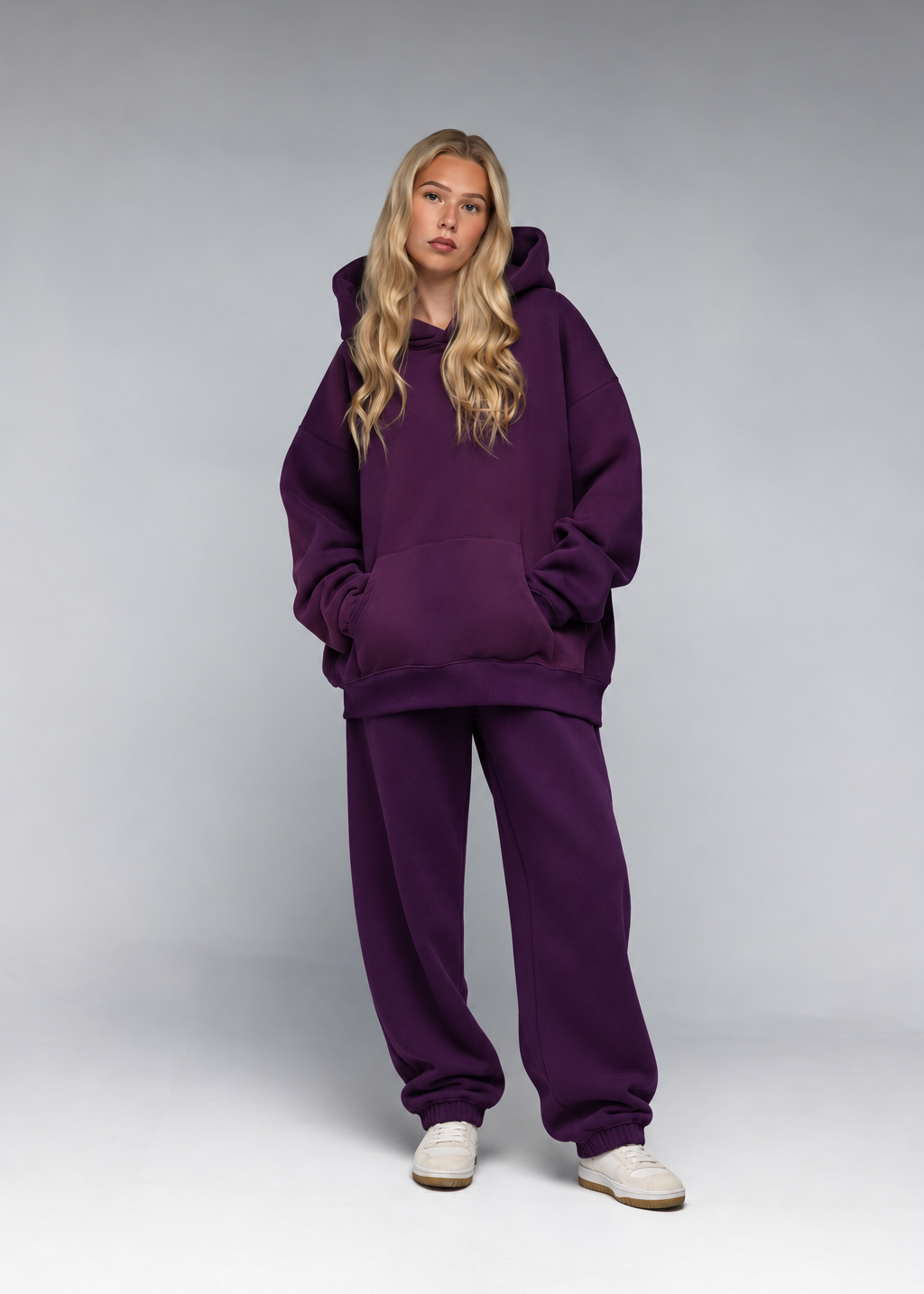 A blonde model wearing an oversized deep purple hoodie and matching sweatpants, standing against a neutral gray studio background.