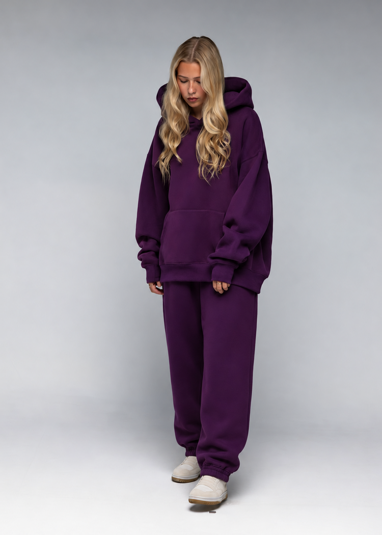 A woman modeling a matching deep purple sweatshirt and sweatpants lounge set, standing against a neutral gray studio background.