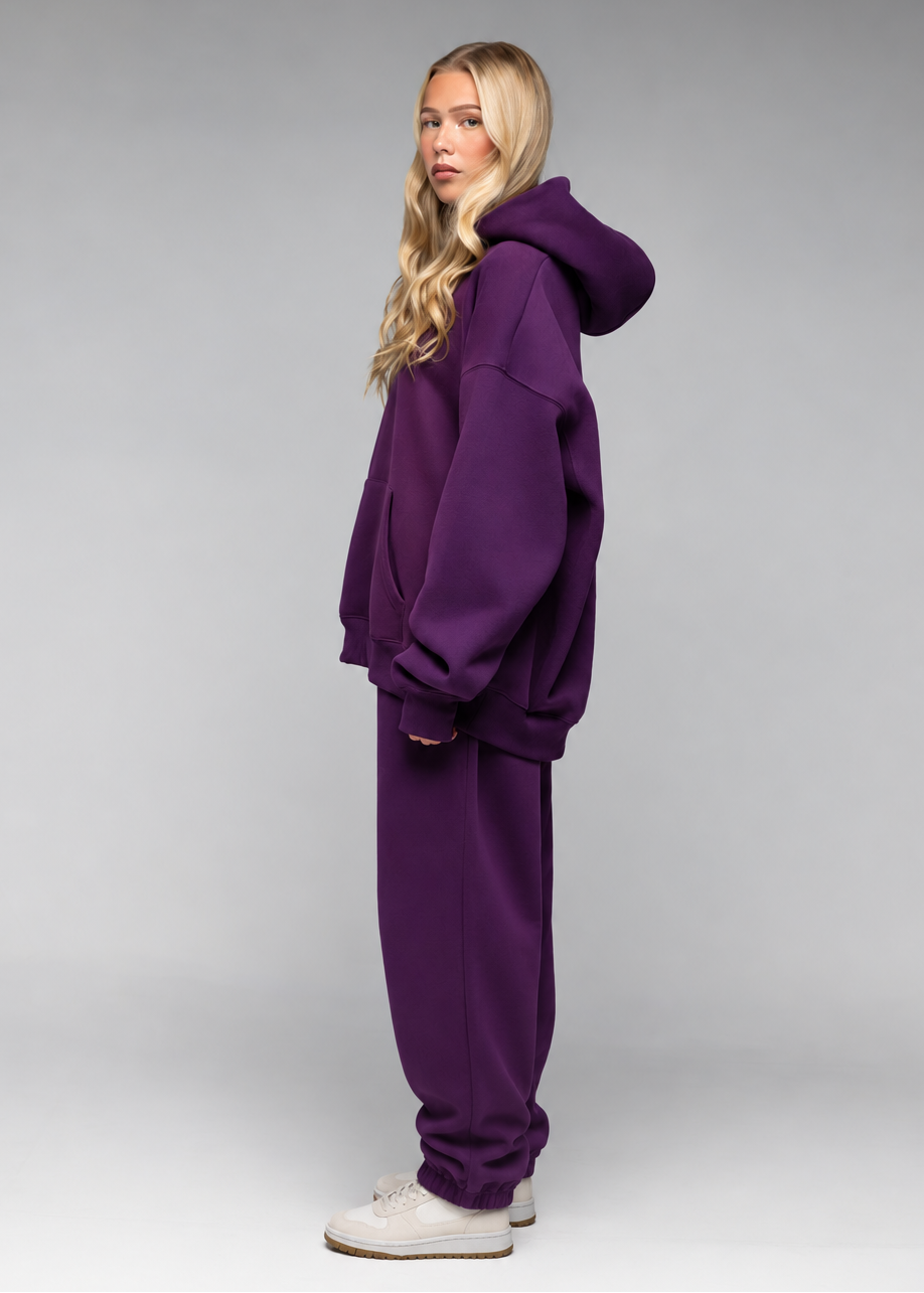 A woman modeling a matching purple sweatshirt and sweatpants lounge set, standing in profile against a neutral gray studio background.
