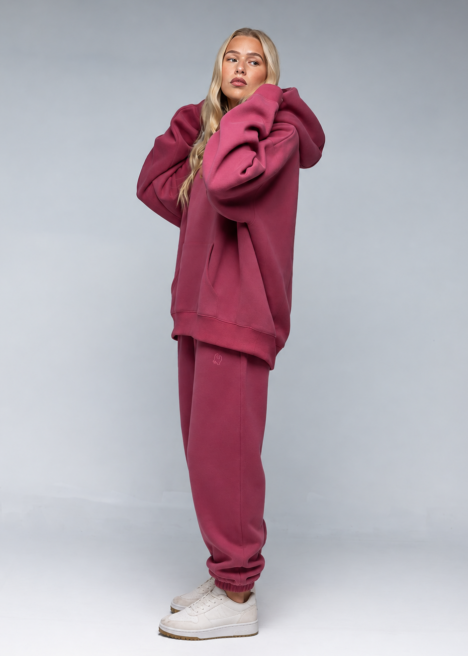 A woman modeling a matching dusty rose sweatshirt and jogger pants set, paired with white sneakers, against a light gray studio background.