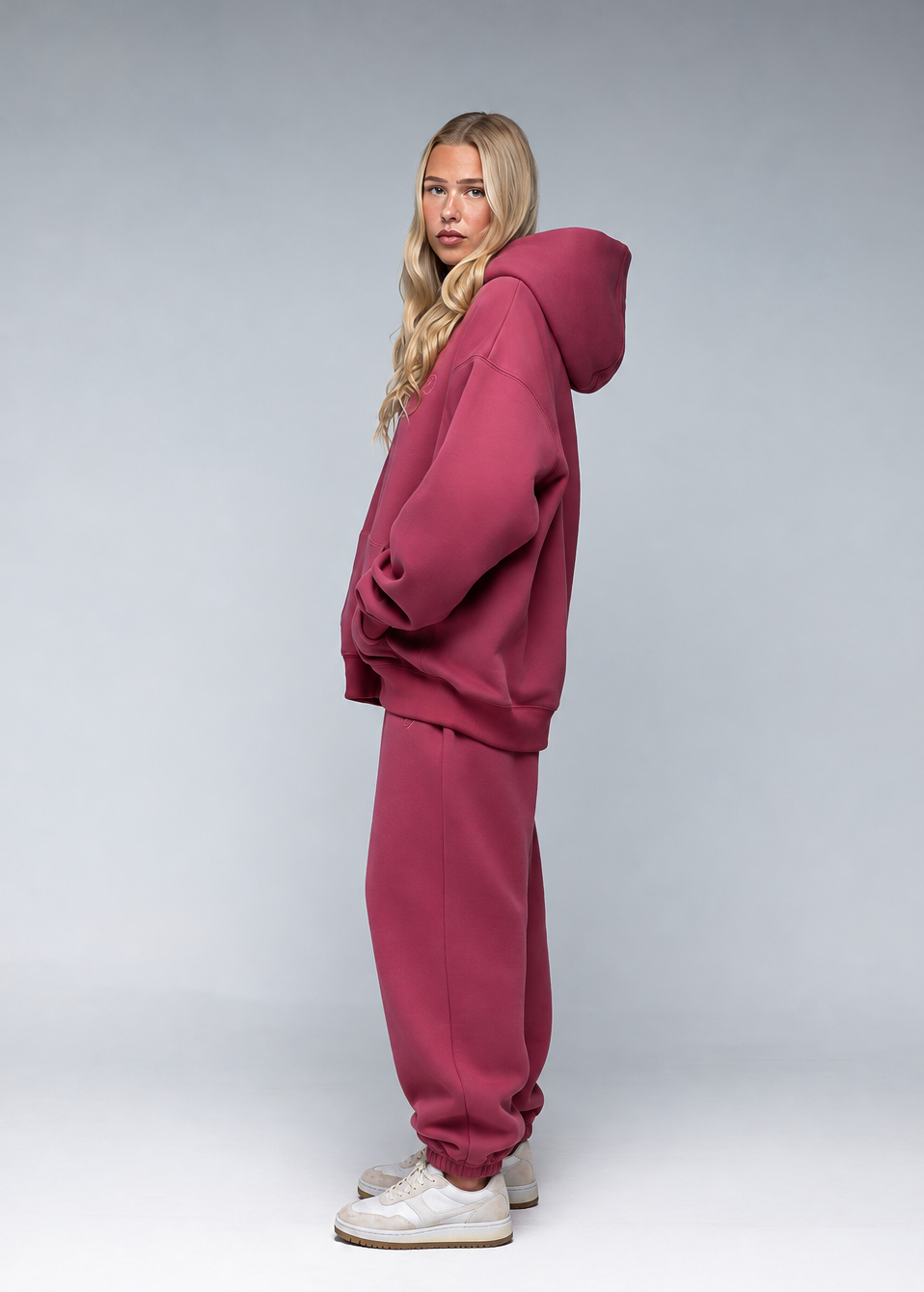 A woman modeling an oversized dusty rose pink tracksuit, consisting of a pullover hoodie and matching jogger pants, paired with white sneakers, set against a neutral gray studio background.