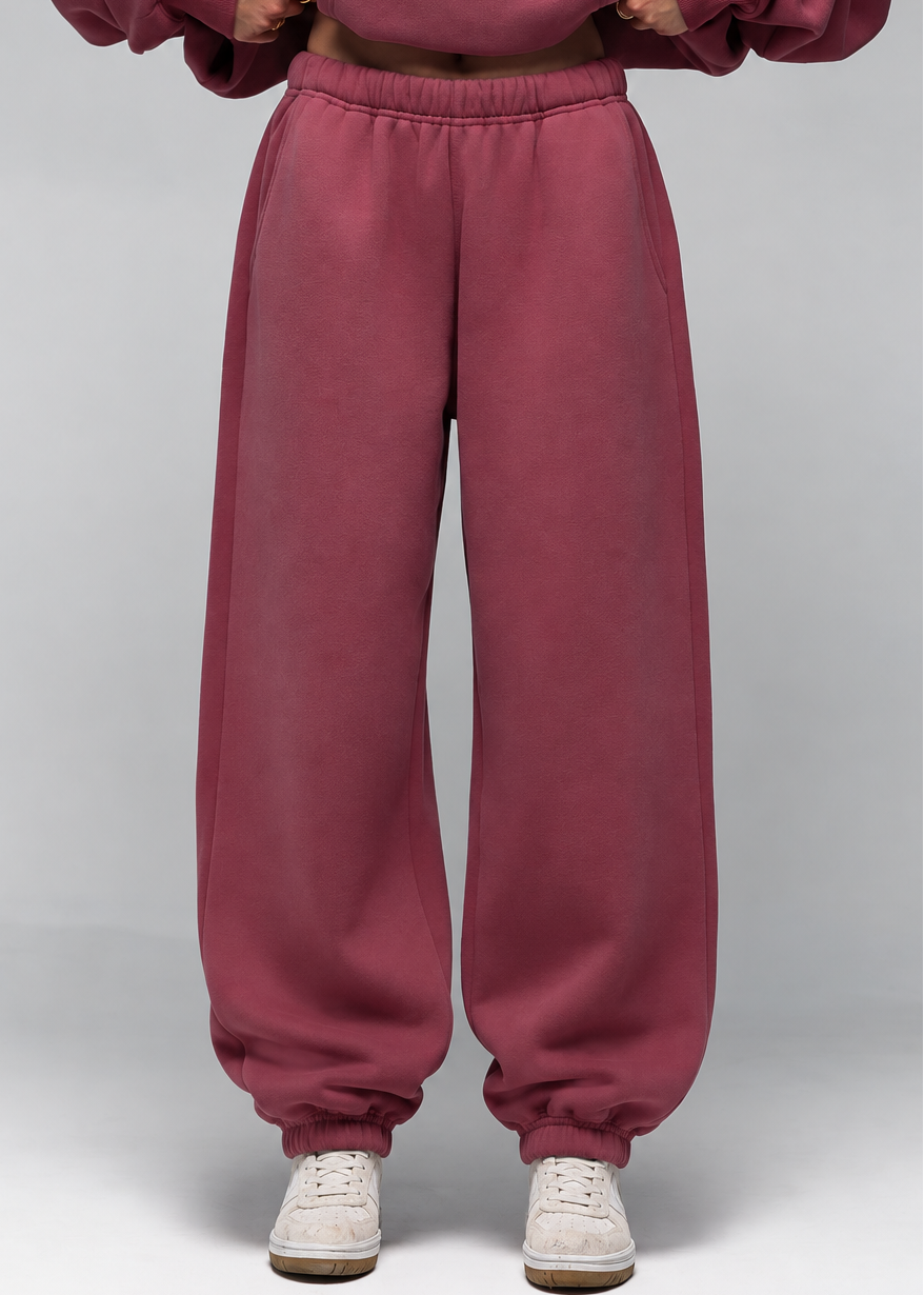 A model wearing a matching dusty rose sweatshirt and sweatpants set, paired with white sneakers, shown from the neck down against a neutral gray studio background.
