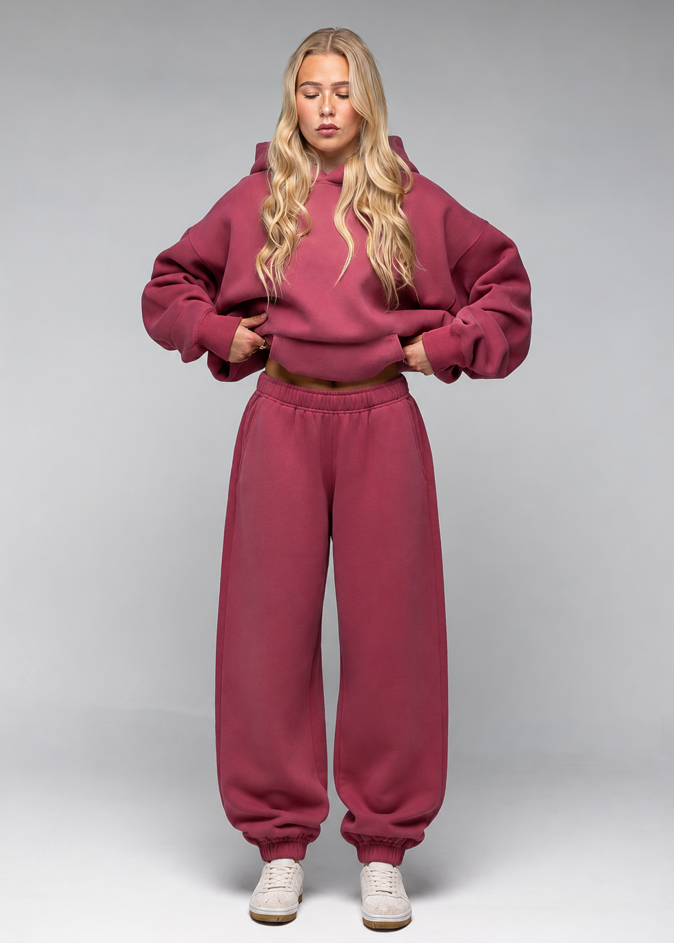 A blonde model wearing a matching dusty rose pink sweatshirt and sweatpants set, standing against a neutral gray studio background.