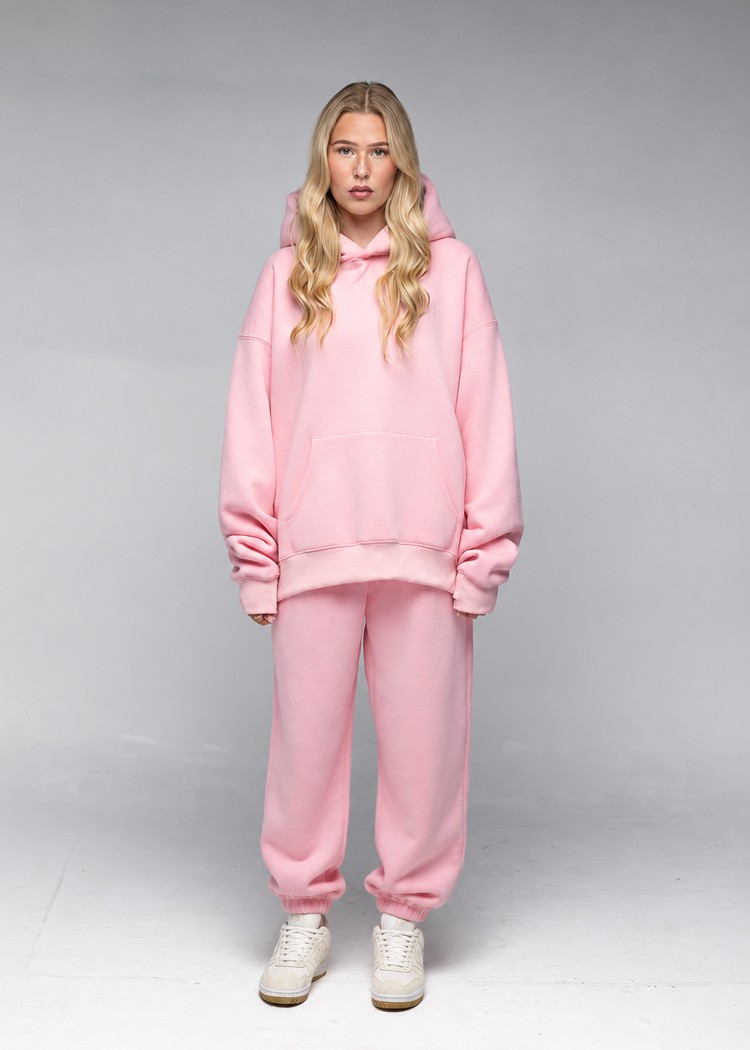 A woman modeling a matching light pink lounge set consisting of an oversized pullover hoodie and jogger sweatpants, paired with white sneakers, against a neutral gray studio background.