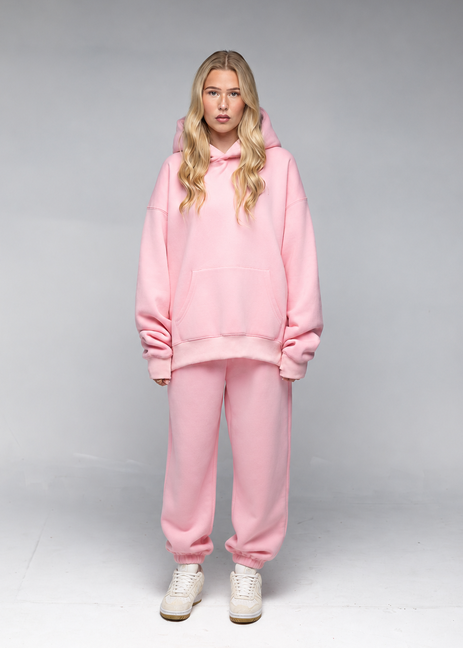 A woman modeling a matching light pink lounge set consisting of an oversized pullover hoodie and jogger sweatpants, paired with white sneakers, against a neutral gray studio background.