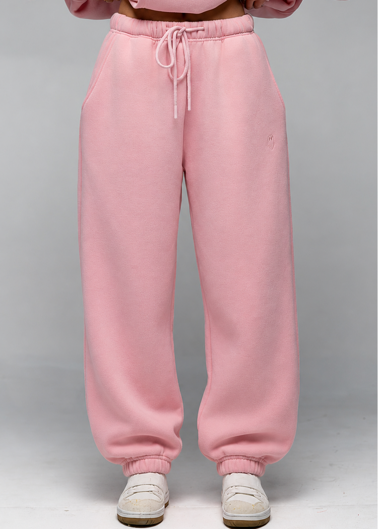 A person wearing loose pink sweatpants with a drawstring waist and white sneakers, shown from the chest down against a light gray studio background.