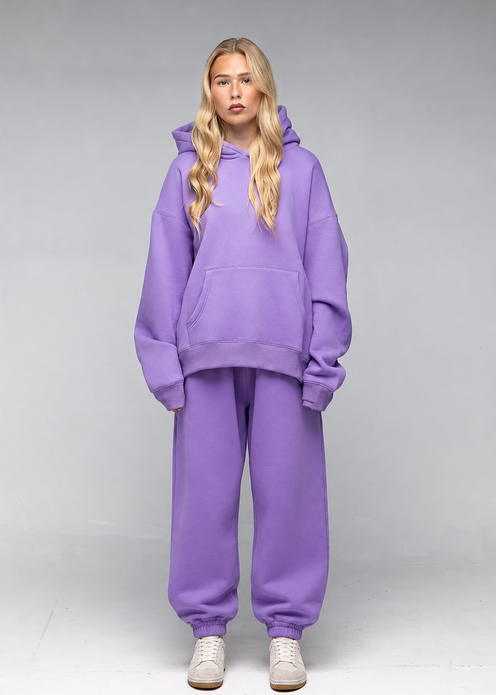 A blonde model wearing an oversized matching purple hoodie and sweatpants set, paired with white sneakers, standing against a neutral gray studio background.