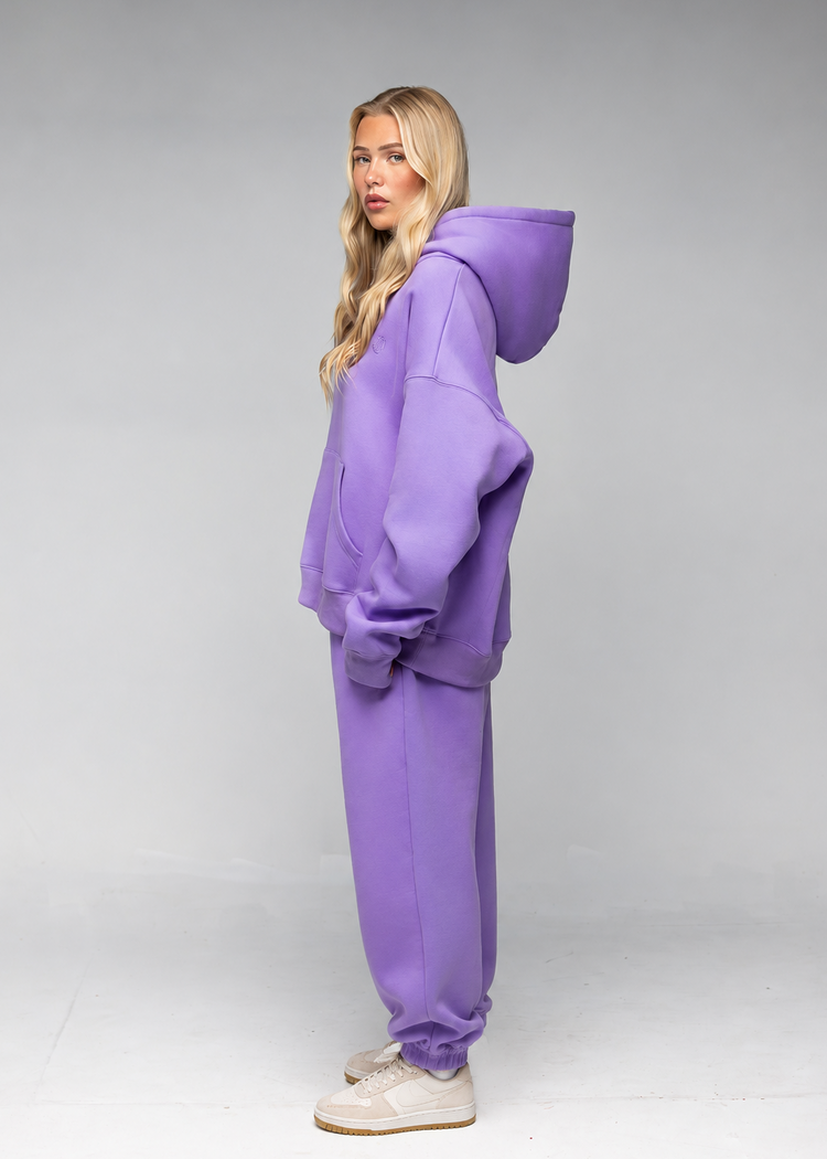 A woman modeling an oversized matching purple hoodie and sweatpants set, standing in profile against a neutral gray studio background.