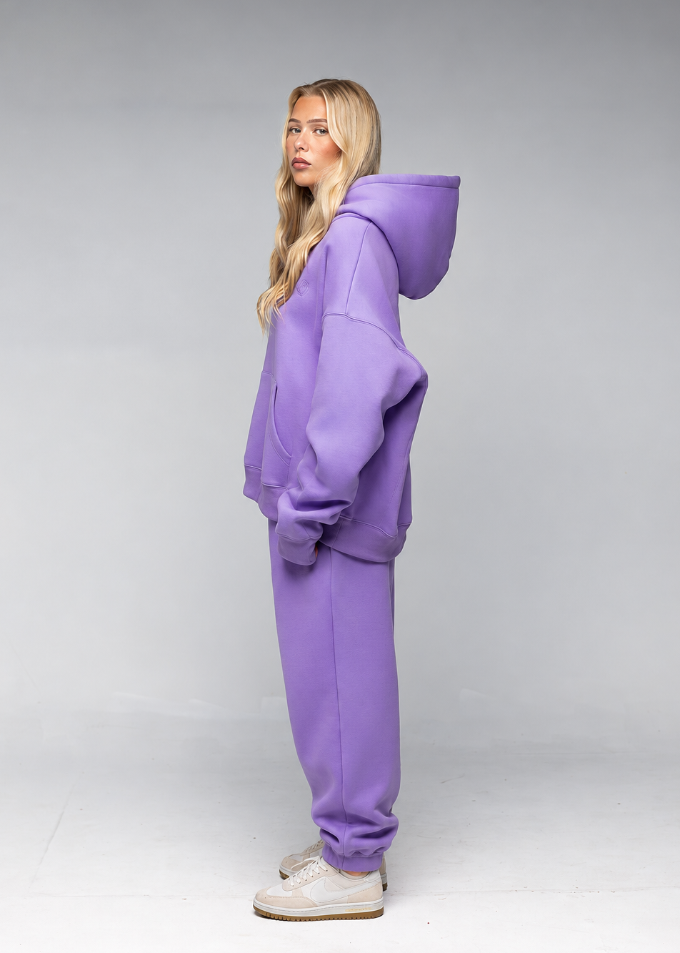 A woman modeling a matching purple lounge set consisting of an oversized zip-up hoodie and sweatpants, paired with white sneakers, against a gray studio background.