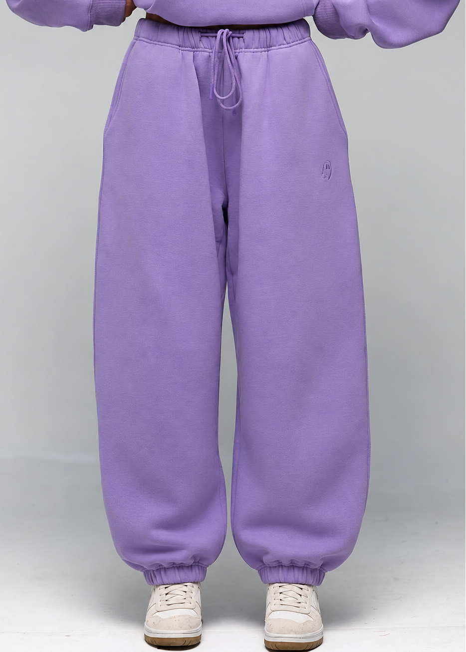 A person wearing loose lavender sweatpants with a drawstring waist and a matching sweatshirt, paired with white sneakers, shown from the chest down against a light gray studio background.