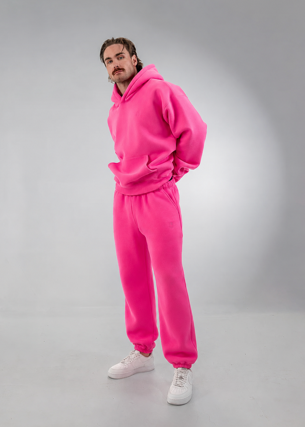 A man modeling a matching hot pink fleece tracksuit, consisting of a pullover hoodie and jogger pants, paired with white sneakers, against a neutral gray studio background.