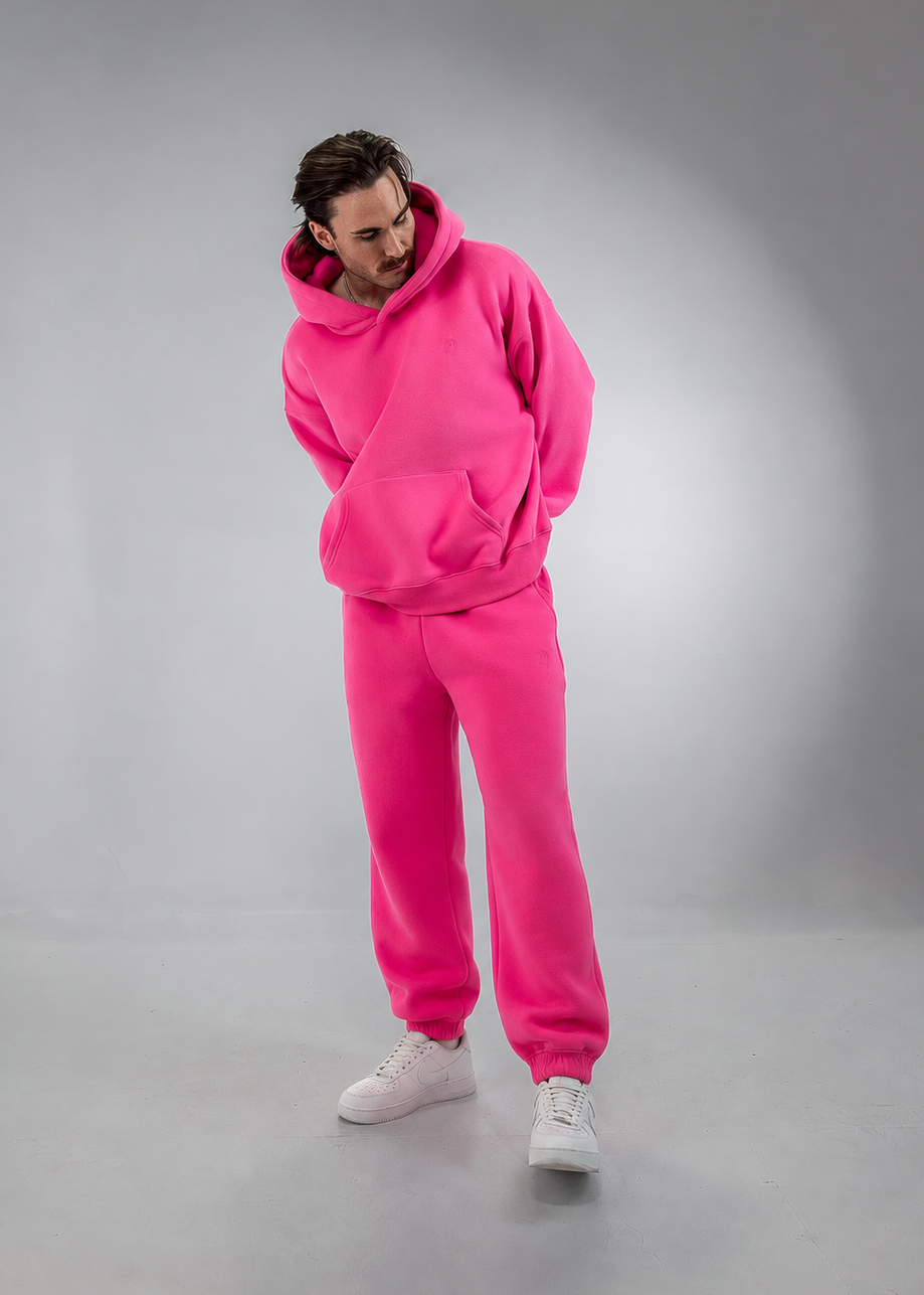A man modeling a matching hot pink fleece tracksuit, consisting of a pullover hoodie and jogger sweatpants, paired with white sneakers, set against a gray studio backdrop.