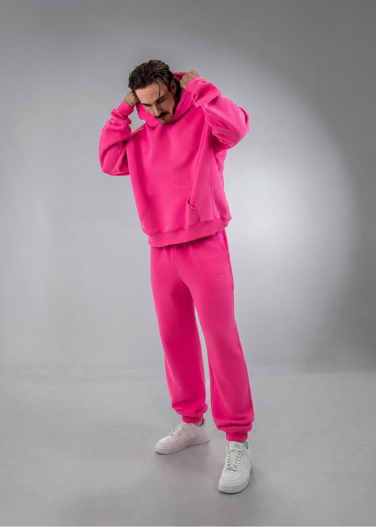 A man modeling a bright pink two-piece tracksuit consisting of a pullover hoodie and matching jogger pants, paired with white sneakers, against a gray studio background.
