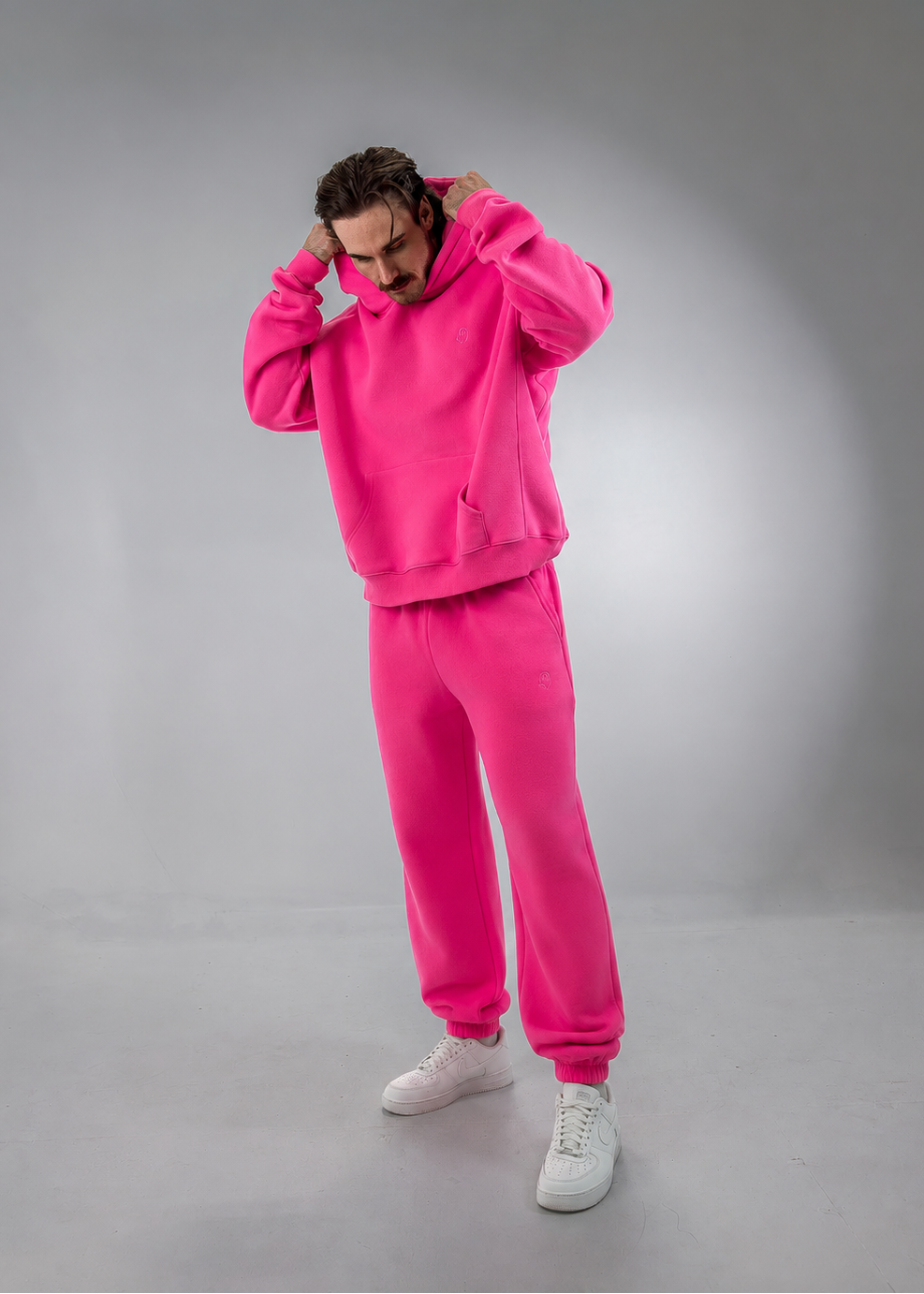 A man modeling a bright pink two-piece tracksuit consisting of a pullover hoodie and matching jogger pants, paired with white sneakers, against a gray studio background.
