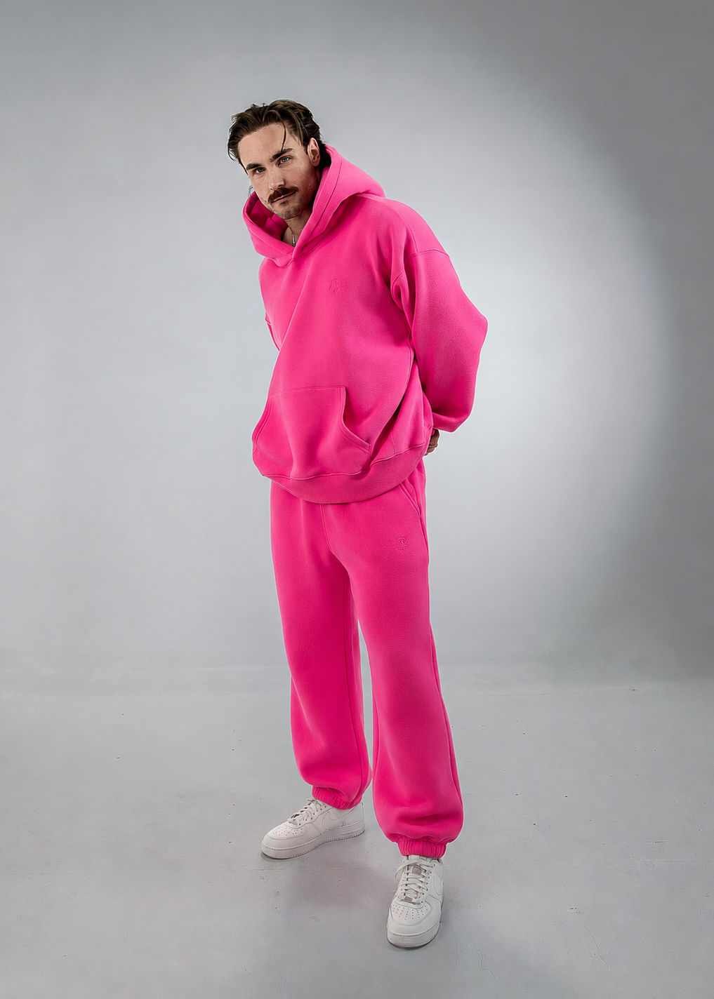 A man modeling a bright pink matching lounge set consisting of an oversized hoodie and sweatpants, paired with white sneakers, against a gray studio background.